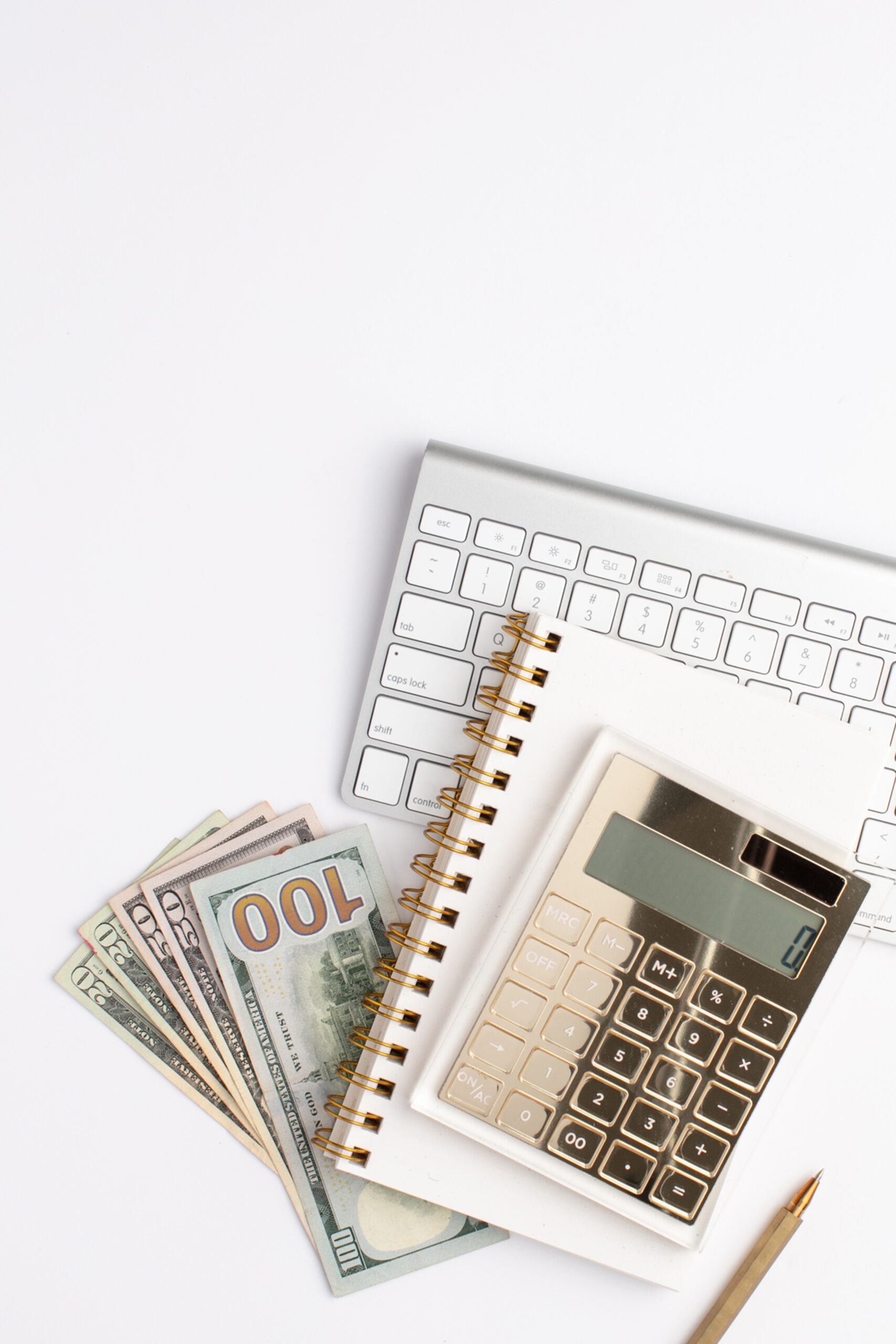 A gold calculator, notebook, keyboard, and hundred dollar bills sit on a white desktop.